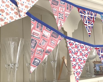 May include: A fabric bunting banner featuring triangular pennants with various British-themed prints, including Union Jack hearts, postage stamp motifs with red backgrounds, and classic Union Jack flags, all attached to a solid blue fabric ribbon.
