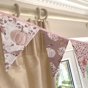 May include: A fabric bunting banner featuring triangular flags with a pink and white colour palette. The patterns include illustrations of pumpkins, leopard print, butterflies, and floral motifs, all connected by a light pink fabric trim.