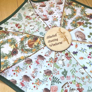 Puede incluir: Una guirnalda de tela con banderines triangulares que presentan diversos estampados de temática forestal, incluyendo conejos, erizos, setas, girasoles y bayas. Una etiqueta circular de madera muestra el texto Bluebell Bunting Wimborne.