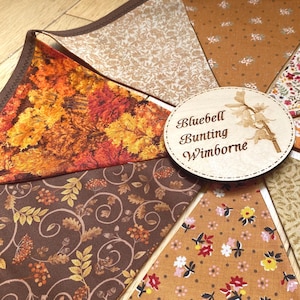 Könnte beinhalten: Eine Stoffwimpelkette mit dreieckigen Flaggen in verschiedenen herbstlichen Mustern, darunter Herbstlaub, braune Eichenblätter und Blumenmotive. Ein rundes Holzschild ist mit dem Text Bluebell Bunting Wimborne bedruckt.