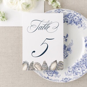 May include: A white table number card with the number 5 and the word "Table" in elegant script, adorned with seashell illustrations. A blue and white floral plate and an envelope overflowing with white and blue flowers complete the scene.