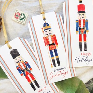 May include: Gift tags with watercolor illustrations of nutcracker soldiers in red, blue, and gold uniforms. Each tag has a gold ribbon and features holiday greetings like "Happy Holidays" and "Season's Greetings" on a striped background.