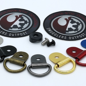May include: A set of seven D-ring hardware pieces in various finishes, including silver, black, brass, gunmetal, gold, red, and blue. The hardware is arranged on a white surface. Two round stickers with the text "Smugglers Outpost" are also included in the image.