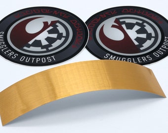 May include: Two round stickers with a black and gray background. The stickers feature a red and white symbol with the text "Smugglers Outpost" in white lettering. The stickers are on a white surface next to a gold metal strip.