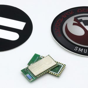 May include: Two circular stickers and a small green electronic circuit board module. One sticker is black with two white horizontal bars. The other sticker features a red and black circular emblem with the text SMUGGLERS OUTPOST in white.