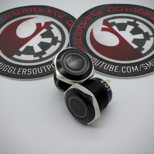 May include: Two black push button switches with silver bezels and a black ring around the button. The switches are on a white surface with two round stickers featuring a Star Wars themed logo.