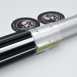 May include: A silver and black lightsaber prop with a detailed design. The hilt features black grips, silver accents, and a red button. Two circular patches with logos are in the background.