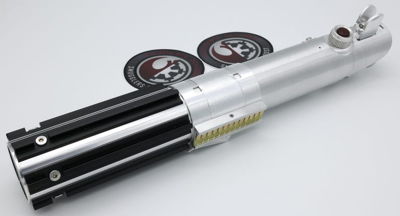May include: A silver and black lightsaber prop with a detailed design. The hilt features black grips, silver accents, and a red button. Two circular patches with logos are in the background.