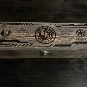 May include: A wooden box with a latch, featuring a Star Wars themed design. The box is engraved with the Rebel Alliance symbol, the Imperial symbol, and the text "Smugglers Outpost".