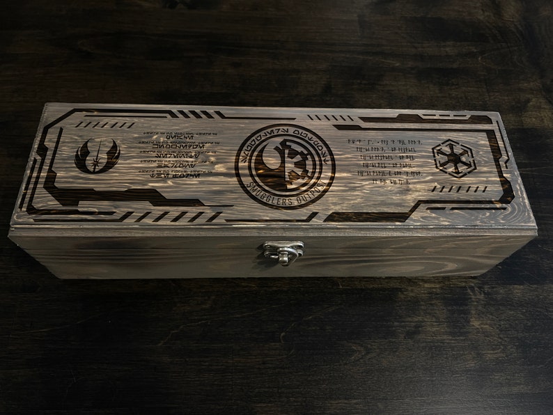 May include: A wooden box with a latch, featuring a Star Wars themed design. The box is engraved with the Rebel Alliance symbol, the Imperial symbol, and the text "Smugglers Outpost".