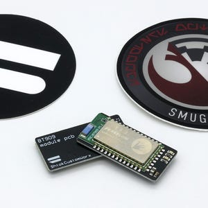 May include: An assortment of electronic components and stickers. Includes a black oval sticker with white shapes, a round sticker with a Rebel Alliance symbol and text, and two circuit boards. One circuit board has the text "BT909 module pcb".