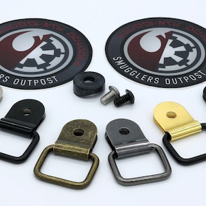 May include: Five different colored metal D-rings with a screw and washer. The D-rings are silver, black, brass, gunmetal, and gold. Two round stickers with the text "Smugglers Outpost" and a Rebel Alliance symbol are in the background.