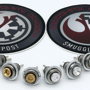May include: A collection of six silver-colored metal push button switches with various colored button tops, including gold and white. Two circular stickers with a red and white emblem and the text "SMUGGLERS OUTPOST" are also visible.