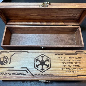 May include: A wooden box with a hinged lid and a brass latch. The box is decorated with a Star Wars Imperial symbol and the text "The Empire Strikes Back".
