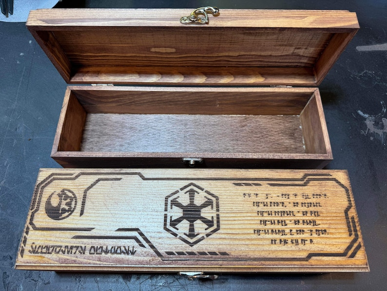 May include: A wooden box with a hinged lid and a brass latch. The box is decorated with a Star Wars Imperial symbol and the text "The Empire Strikes Back".