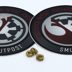 May include: Two circular black and red stickers with the text "SMUGGLERS OUTPOST" and a graphic design. Three small brass nuts are also in the image. The stickers have a Star Wars theme.
