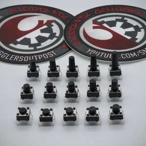 Puede incluir: Una colección de 16 interruptores táctiles de botón pulsador negros con carcasas de metal plateado. Los interruptores están dispuestos en cuatro filas de cuatro. Los interruptores están sobre una superficie blanca.