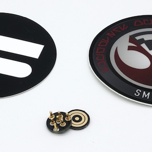 May include: Two circular adhesive decals and a small electronic component with gold pins. One decal is black with two white horizontal bars. The other is a circular emblem featuring a red and silver design with the text SMUGGLERS OUTPOST.