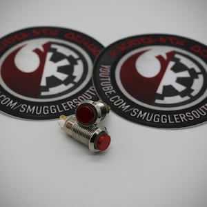 May include: Two silver and red push button switches with the text "YOUTUBE.COM/SMUGGLERSOUTPOST" on two black and white stickers with the Rebel Alliance and Imperial symbol.
