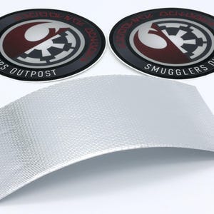 May include: Two circular black, red, and silver Smugglers Outpost patches with a curved silver textured material. The patches feature a stylized design with text around the edge. The silver material has a woven pattern.
