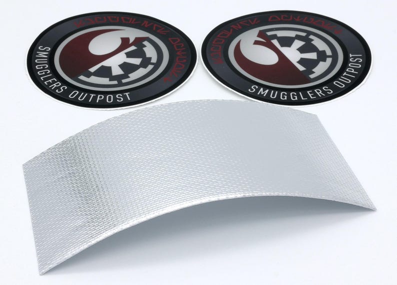 May include: Two circular black, red, and silver Smugglers Outpost patches with a curved silver textured material. The patches feature a stylized design with text around the edge. The silver material has a woven pattern.