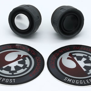Peut inclure: Deux haut-parleurs cylindriques noirs avec des centres argentés et des côtés striés sont affichés au-dessus de deux écussons circulaires. Les écussons présentent un emblème rouge et blanc avec le texte "SMUGGLERS OUTPOST" et d'autres textes dans une police stylisée.