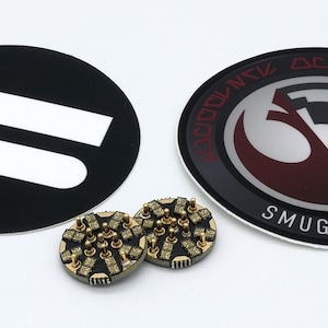 May include: Two circular stickers and two small round electronic components. One sticker is black with two white horizontal bars. The other is black with a red and silver emblem and the text SMUGGLERS OUTPOST. The components have gold pins.