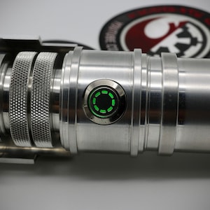 May include: Close-up of a silver lightsaber hilt with textured sections and a green illuminated power button. The design is detailed and futuristic, suggesting a sci-fi theme. The hilt is made of metal.