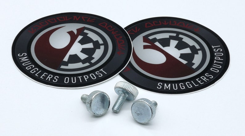 K&ouml;nnte beinhalten: Zwei runde schwarze und silberne Aufkleber mit dem Text "Smugglers Outpost" und einem Rebellenallianz-Symbol und einem imperialen Symbol. Vier silberne R&auml;ndelschrauben.