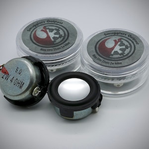May include: Two small, silver speakers with black rims and the text "2W 4 OHM". Two clear plastic containers with gray lids are also present. The lids have the text "Smugglers Outpost" and "Nano Elite 22mm 2w 4ohm".