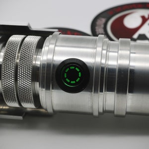 May include: Close-up of a silver lightsaber hilt with a green LED light. The hilt has a textured surface and a black button with a circular design.