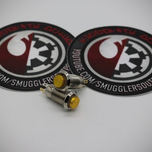 May include: Three gold-colored push button switches with silver metal housings. The switches are on a white surface with two round stickers featuring a Rebel Alliance symbol and the text "YOUTUBE.COM/SMUGGLERSOUTPOST".