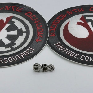 May include: Two round black and white stickers featuring the Rebel Alliance and Imperial logos, alongside the text "YOUTUBE.COM/SMUGGLERSOUTPOST". Four silver screws are also shown.