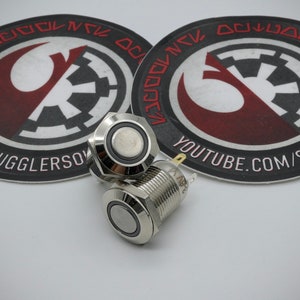 May include: Two silver push button switches with a black ring around the button. The switches are on a white surface with two round stickers featuring the Star Wars Rebel Alliance and Imperial symbols.