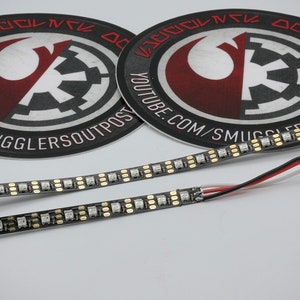 May include: Two black and white LED light strips with wires attached. The strips are on a white surface. The image also shows two round stickers with the text "YOUTUBE.COM/SMUGGLERSOUTPOST" and a Rebel Alliance symbol.