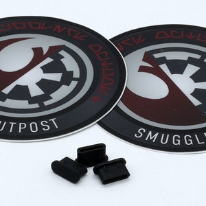 May include: Two round black and gray stickers with the text "Smugglers Outpost" and a Rebel Alliance symbol with an Imperial symbol superimposed on it. Four black rubber plugs are also in the image.