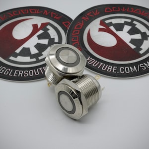 May include: Two silver push button switches with a black ring around the button. The switches are stacked on top of each other. The switches are on a white surface.