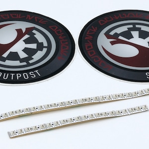 May include: Two round stickers with a black and white imperial symbol and a red and white rebel symbol, with the text "Smugglers Outpost" around the edge. Three white LED light strips are also shown.