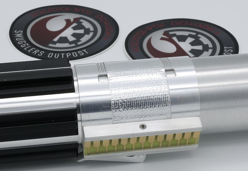 May include: A silver lightsaber hilt with black accents and a rectangular section with gold-colored connectors. Two circular patches with the text "Smugglers Outpost" and a logo are in the background.