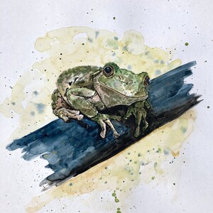 May include: A watercolor painting of a green frog sitting on a blue branch. The frog is facing the right side of the image and has a light green body with darker green markings. The background is a light yellow with green and brown splatters.