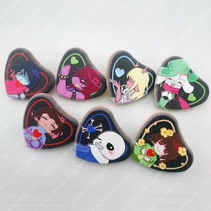 May include: Seven heart-shaped pins featuring colorful anime-style character illustrations on a black background. Each pin displays a unique character, including figures with red, pink, yellow, and blue accents, set within distinct colored heart outlines.