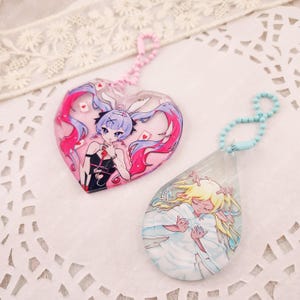 May include: Two acrylic keychains with ball chains. One is a heart-shaped charm featuring an anime character with long pink and blue hair, bunny ears, and playing cards. The other is a teardrop-shaped charm with a character with blonde hair.