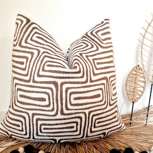 Puede incluir: Una almohada de tiro de forma cuadrada con un patrón geométrico blanco y marrón. La almohada tiene un patrón geométrico marrón sobre un fondo blanco.