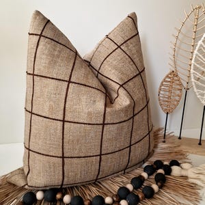May include: A large beige pillow with a dark brown grid pattern. Decorative wooden leaf accents and a beaded garland with black, white, and natural wood beads are in the foreground. The pillow is on a woven mat.