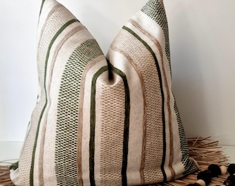 May include: A decorative pillow with a striped pattern in shades of beige, green, and brown. The pillow has a textured appearance and is placed on a woven mat with decorative wooden beads. The pillow's design features vertical stripes.