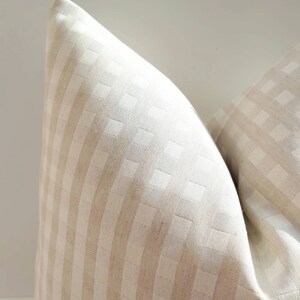May include: A close-up of a cream-colored pillow with a subtle checkered pattern. The pillow is made of a soft, textured fabric.