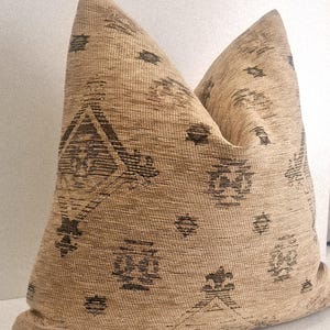 Puede incluir: Cojín decorativo beige con textura tejida y patrones geométricos marrón oscuro. La almohada tiene un aspecto suave y acogedor, adecuado para añadir un toque de estilo a cualquier espacio habitable.