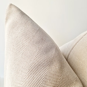 May include: Two beige throw pillows with a subtle herringbone pattern. The pillows are made of a soft, woven fabric.