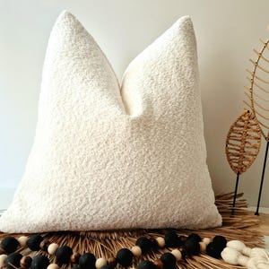 May include: A large, cream-coloured textured cushion with a soft, plush appearance. The cushion is placed on a woven mat with decorative black and white beads. The cushion's neutral colour and texture suggest a cosy, modern aesthetic.