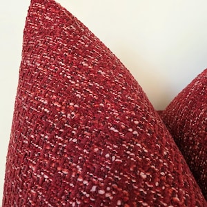 May include: A textured throw cushion cover featuring a deep red and burgundy woven fabric with subtle flecks of white and light pink throughout. The material has a thick, multi-dimensional weave that creates a dense, tactile surface appearance.